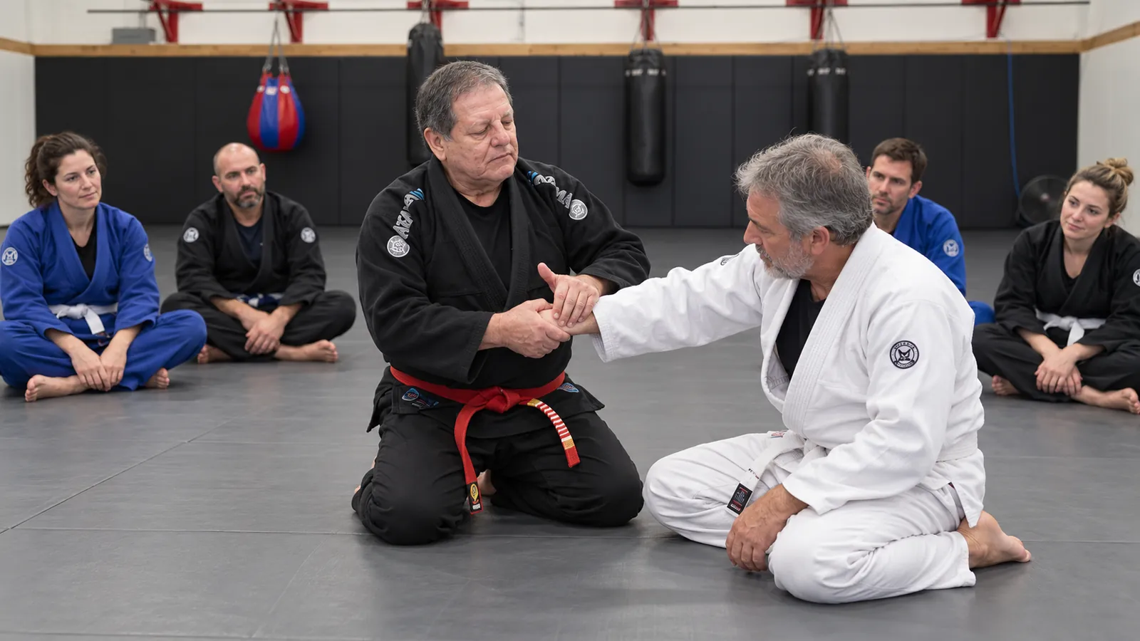 A red-belt instructor demonstrates a wrist grip on an older student as the class watches