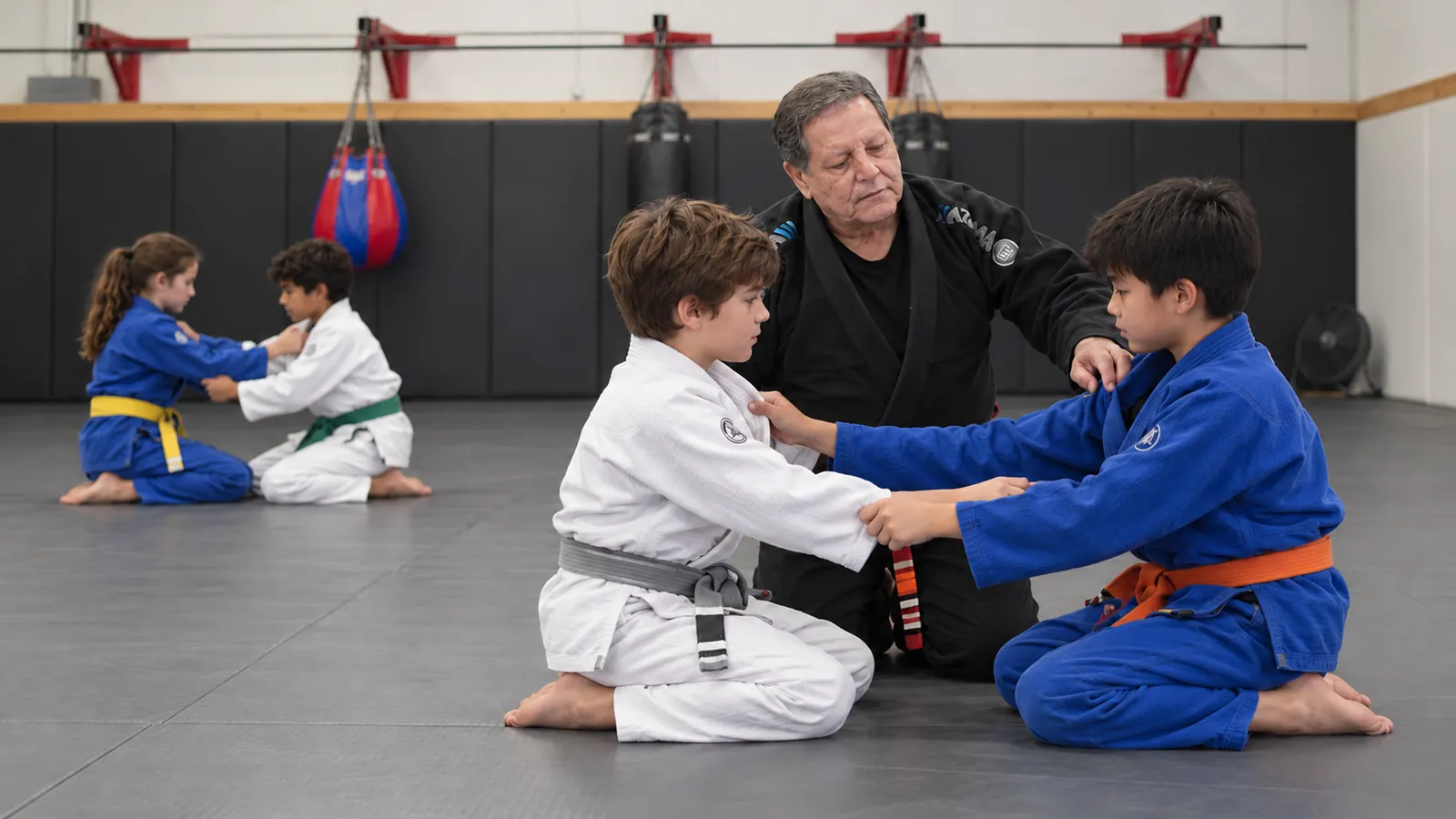 Children in grey, orange, yellow and green belts practise grips in pairs with an instructor